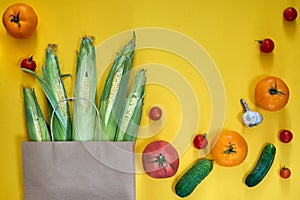 Paper craft bag with different groceries on yellow background. Top view cherry tomatoes, cucumbers, garlic. Corn harvest farm shop