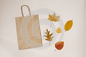 A paper craft bag with shopping handles and autumn fallen leaves yellow and red on a beige background. Autumn discounts, price fal
