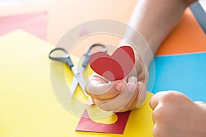 Paper crafts for kids. Child hands cuts colored paper with scissors and holding red paper heart