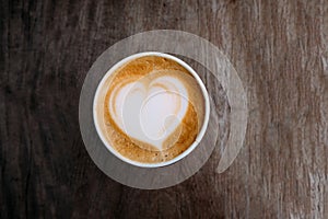 Paper cup of cappuccino coffee with beautiful latte art in the shape of heart on wooden background. Top view, copy space.