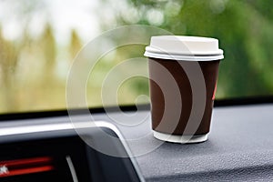 Paper cup with coffee on dashboard of car on road background.