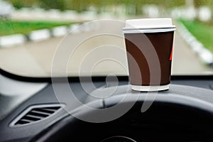 Paper cup with coffee on dashboard of car on road background.