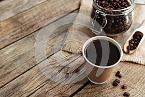 Paper cup of coffee, dry coffee beans in glass jar and spoon at wooden table