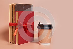 A paper cup of coffee stands next to a stack of old books tied with a red ribbon