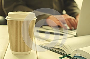 Paper cup of coffee on work-table with office stuff, notepad, laptop, pen, coffee cup and working man on background