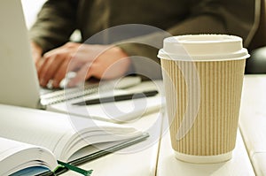 Paper cup of coffee on work-table with office stuff, notepad, laptop, pen, coffee cup and working man on background