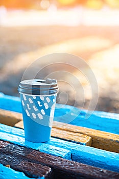 Paper cup with hot drink on wooden painted bench in the sun in the park