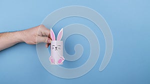 Paper Easter bunny in hand on a blue background. the hand holds the rabbit by the ear