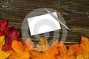 Paper with an empty space for text on a pile of bright autumn leaves against a dark wooden table