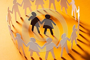 Paper figure of female couple surrounded by circle of paper people holding hands on yellow surface. Bulling, minorities