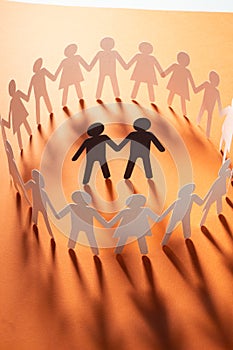 Paper figure of male couple surrounded by circle of paper people holding hands on orange surface. Bulling, minorities
