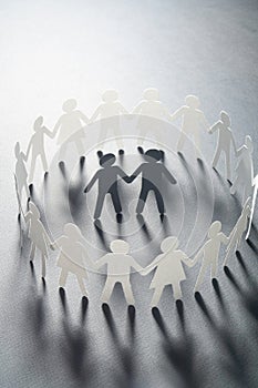Paper figure of a male couple surrounded by circle of paper people holding hands on white surface. Bulling, minorities