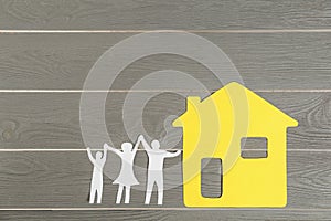 Paper figures of happy family holding  and yellow house on grey wooden background, flat lay