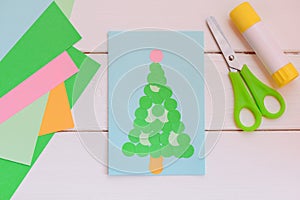 Paper greeting card with Christmas tree, colored paper sheets, scissors, glue stick on a wooden table. Easy paper Christmas tree