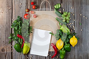 Paper grocery bag with blank notebook and fresh vegetables and fruits on wooden table top view. Eco shopping concept and product