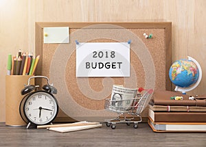 paper note with 2018 Budget word on cork board with alarm clock, book, money on wooden desk.