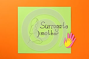 Paper note with drawn pregnant woman figure and words Surrogate mother on orange background, top view