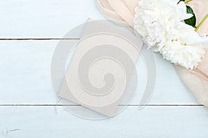 Paper notebook and beige peony flowers on blue wooden table top view. Feminine workspace, flat lay style composition