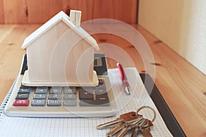 Paper notebook with calculator, house model, keys and pen on wooden table.