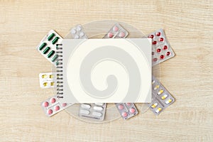 Paper notepad mock up in frame of various blisters with multi-colored tablets and capsules on wooden table. Doctor desk