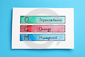 Paper with OCM abbreviation and its interpretation Organizational Change Management on light blue background, top view