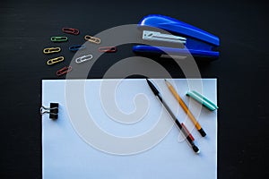 A paper and pencils stepler and colored clips on on black background