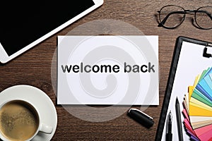 Paper with phrase Welcome Back on wooden table. Office desk with tablet and coffee, flat lay photo