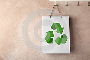 Paper shopping bag with recycling symbol hanging on beige wall. Space for text