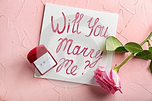 Paper with text WILL YOU MARRY ME? and engagement ring on color background