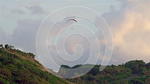 The paraglider soars by parachute on the green mountains. In the distance there is an observation deck with people watching the su