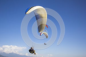 A paraglider on a white parachute in a clear blue sky against the backdrop of mountains and clouds and other pilots
