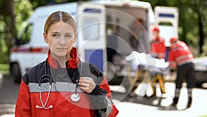 Paramedic posing for camera, ambulance crew transporting patient on background