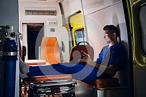 Paramedic in uniform sits with phone in his hands in the ambulance car at night