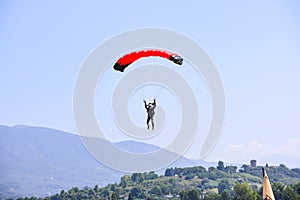paratrooper with red parachute getting ready for landing. On background the blue sky, mountains and green hills