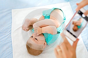 Parent taking photo of a baby with smartphone. Adorable newborn child taking foot in mouth