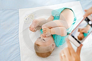 Parent taking photo of a baby with smartphone. Adorable newborn child taking foot in mouth