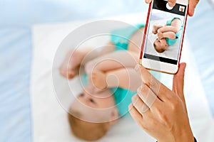 Parent taking photo of a baby with smartphone. Adorable newborn child taking foot in mouth