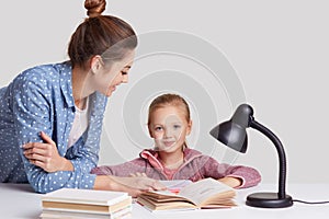 Parenthood, studying and education concept, Attractive blue eyed female child sits at workplace, reads book together with mother,