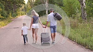Parents with child go with a suitcase in their hands on an empty road in forest.