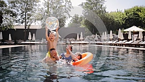 Parents with children playing with ball in swimming pool water in sunny summer. Active family vacation at a beach resort