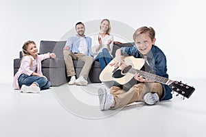 Parents and cute little sister looking at smiling boy playing guitar