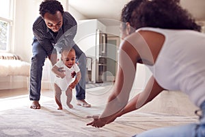 Parents Encouraging Smiling Baby Daughter To Take First Steps And Walk At Home