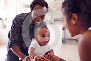 Parents Encouraging Smiling Baby Daughter To Take First Steps And Walk At Home