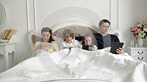 Parents and Kids Using Mobile Devices Together in the Bedroom During Weekend at Home. Young Couple with Children are