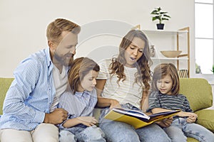 Parents and little children reading book of stories while sitting on couch at home