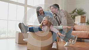 Parents pushing cardboard box with little daughters riding inside, happy family homeowners having fun on moving day