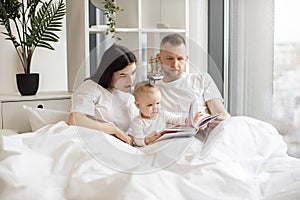 Parents reading baby book with daughter in bed at home