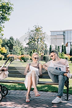 parents sitting on bench near baby carriage in park and reading magazine