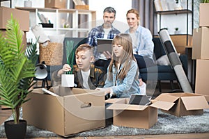 Parents sitting on sofa with laptop, while kids playing and unpacking boxes at new flat