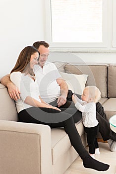 Parents sitting on the sofa and looking at their beautiful baby girl playing with cell phone.